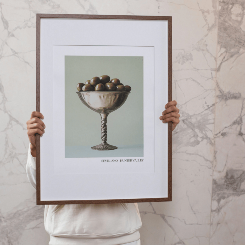 Person holding a framed Sevillano olive poster against a marble wall. 