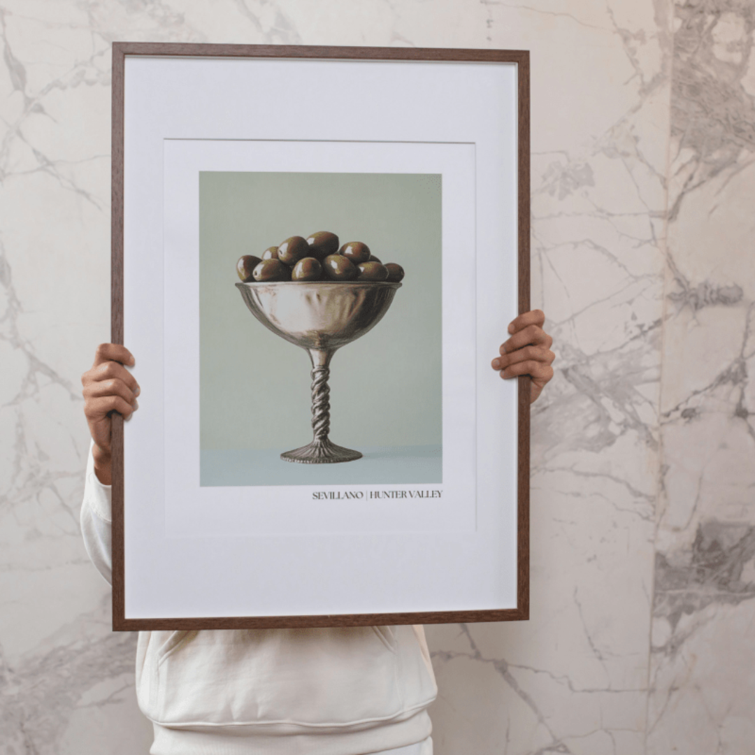 Person holding a framed Sevillano olive poster against a marble wall. 
