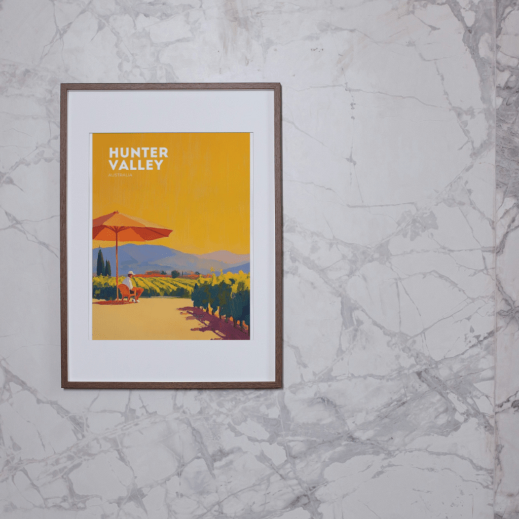 Framed poster on a marble wall of a vibrant Hunter Valley vineyard scene, featuring a person relaxing under a red umbrella against a golden sky.