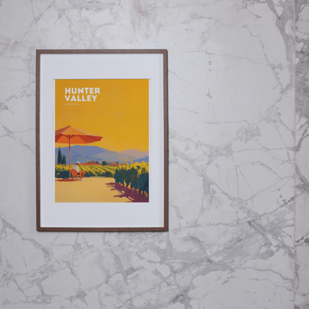 Framed poster on a marble wall of a vibrant Hunter Valley vineyard scene, featuring a person relaxing under a red umbrella against a golden sky.